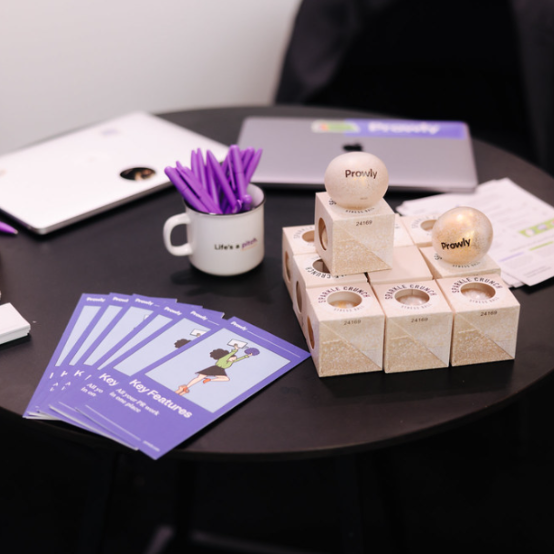 High-quality event merchandise displayed on a table. It includes a branded cup with purple branded pens, branded stressballs in packaging, and leaflets. There is a branded folder in the background.