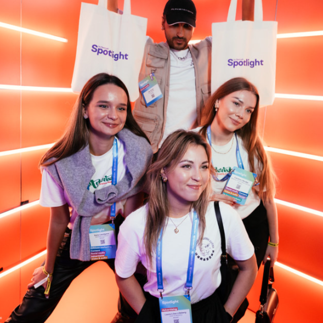 Four people including three smiling women and one man with a baseball cap on. They are wearing high-quality swag including tote bags, visitor tags, and t-shirts with a Spotlight logo on.