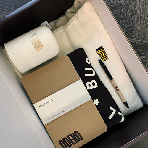 A beautiful box of high-quality swag for Odeko. It consists of branded items including a sweatshirt, cup, notebook, t-shirt, and pen.