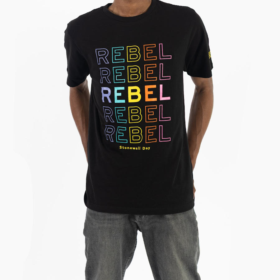 A person wearing a black t-shirt with "REBEL" printed multiple times in a vertical stack of neon purple, orange, and yellow colors, with "Stonewall Day" printed at the bottom. Stonewall Day Merch, Custom Rebel T-shirt, Advocacy Screen Printing, Premium Campaign Swag, TVP NYC Apparel Solutions.