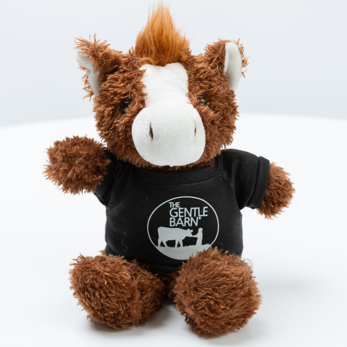 Plush toy cow merch from TVP NYC wearing a black shirt with 'The Gentle Barn' logo on a white background