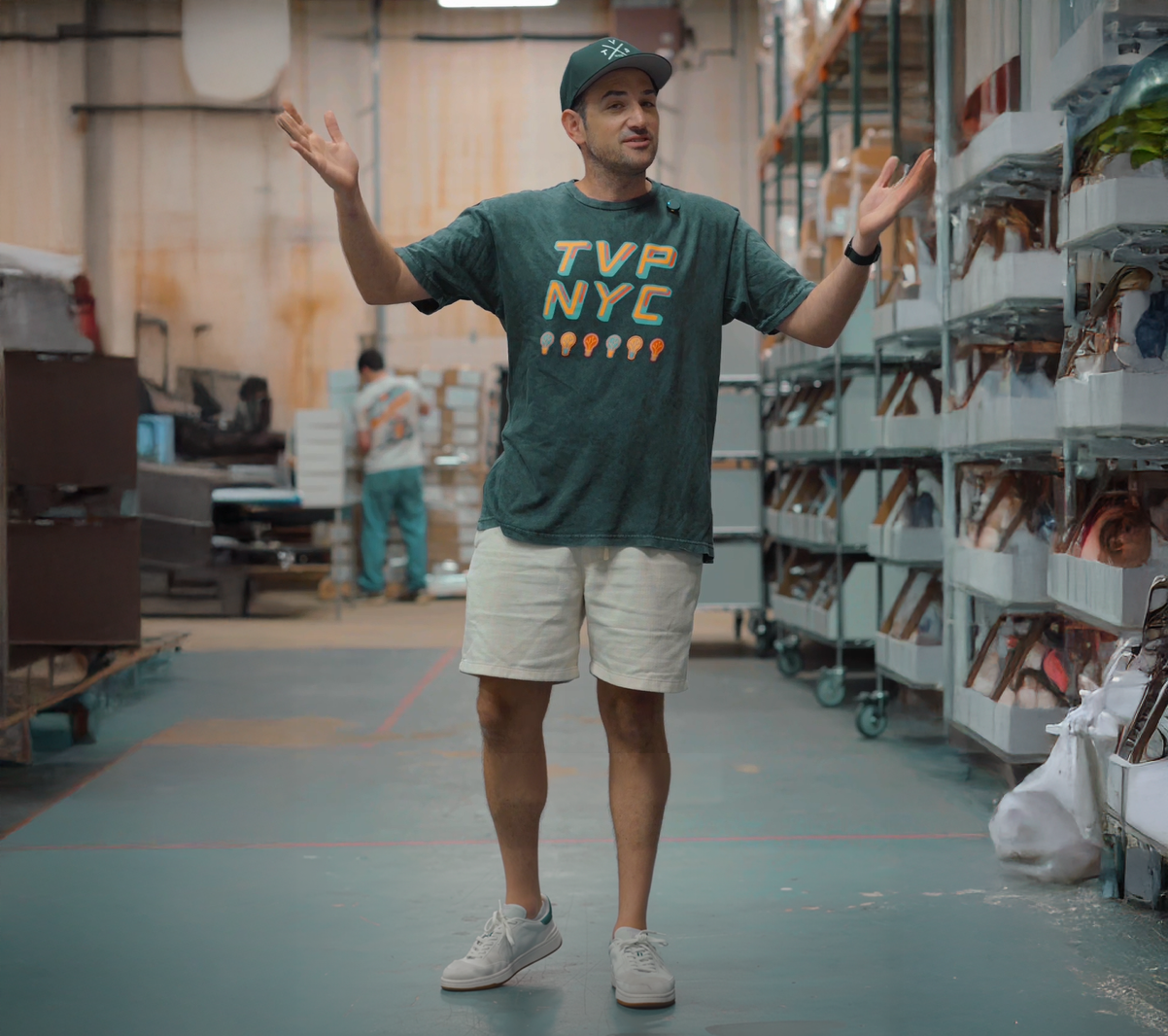 Man in a warehouse wearing a green 'TVP NYC' t-shirt with arms outstretched.