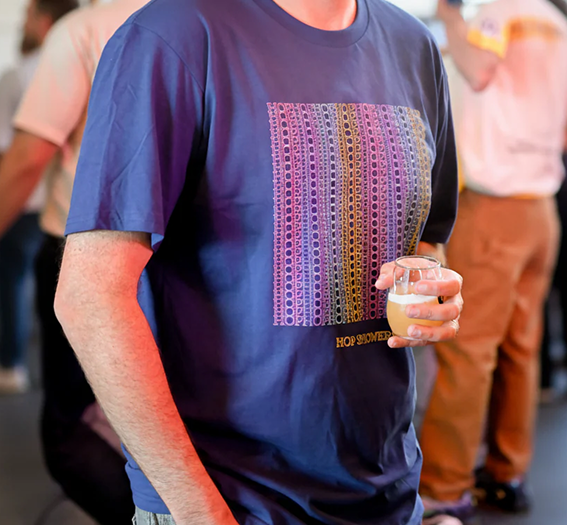 Person wearing a blue t-shirt with a colorful graphic design, holding a glass of beer in a social setting. This image shows cool swag in a social environment.