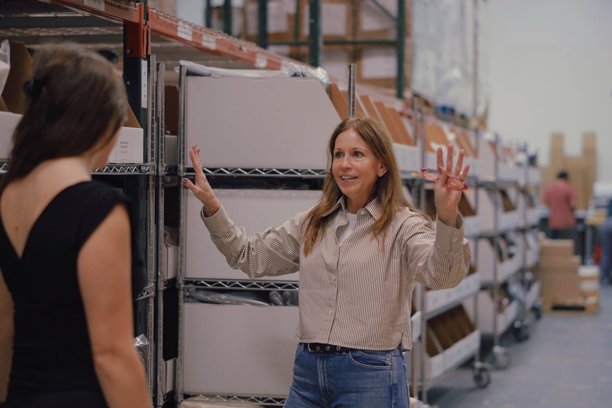 Woman in the TVP NYC smiling logistics coordinator gesturing toward organized warehouse shelving filled with white inventory bins containing packaged apparel and brand merchandise.